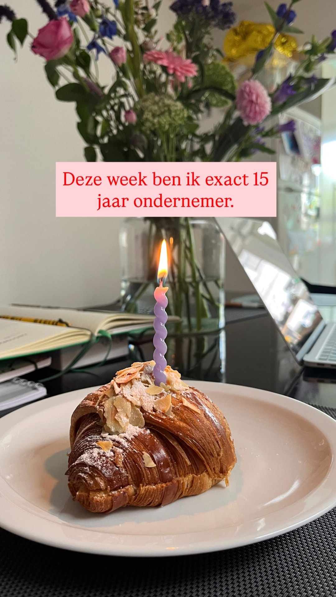 Een amandelcroissant met een brandend kaarsje op een tafel met laptop, notitieboek en een vaas bloemen